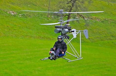 A single-seat helicopter flying over a grassy field.