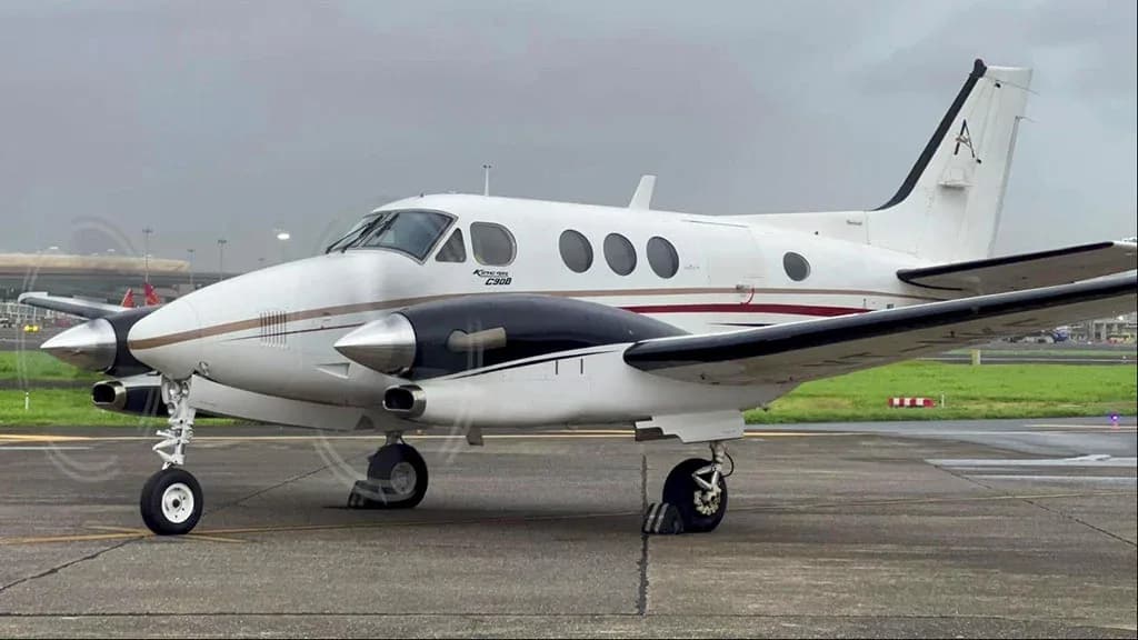 A white Beechcraft King Air C90B with brown and black stripes on the fuselage, parked on an airfield under an overcast sky.