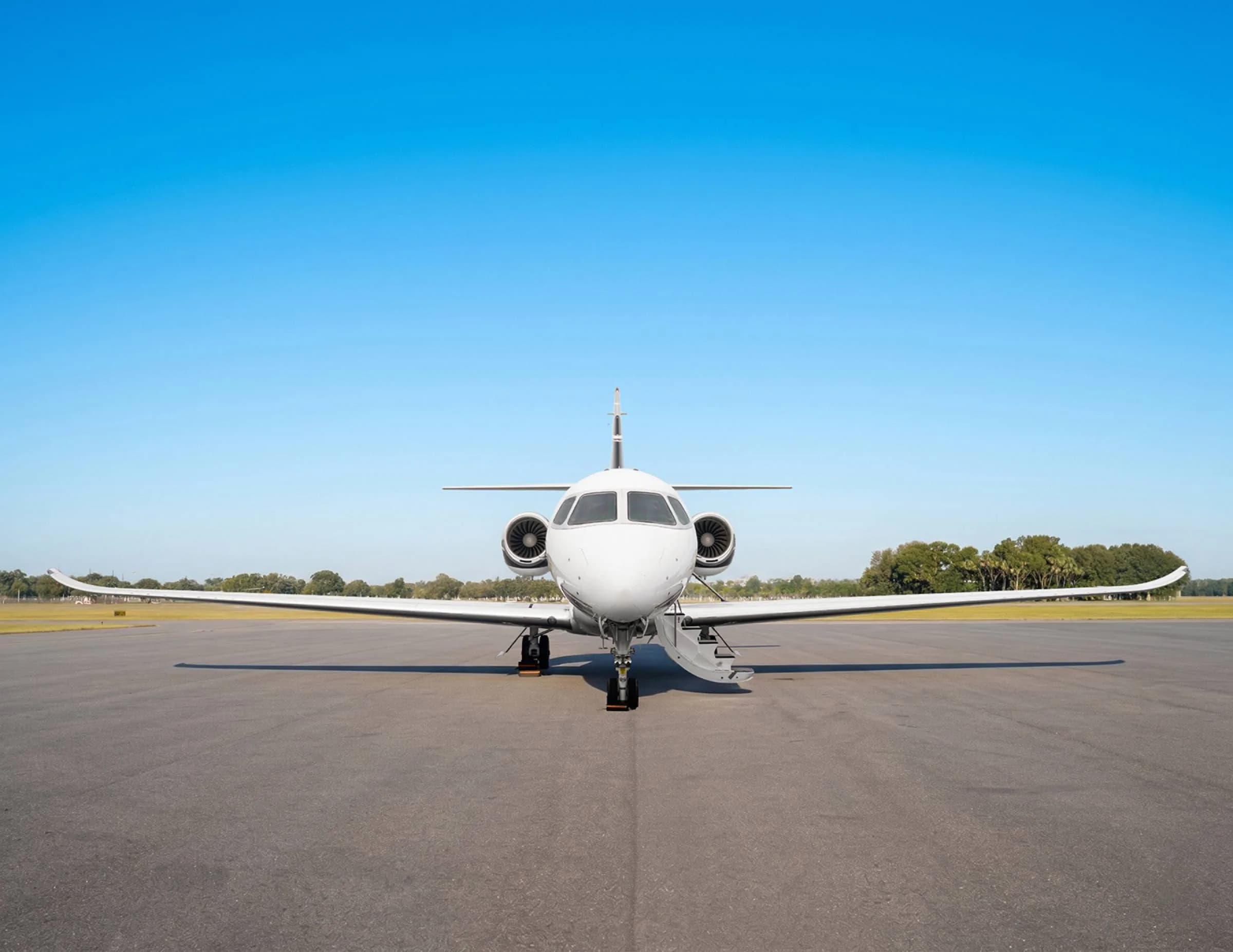 A head-on view of a white private jet with its engines visible, parked on an asphalt runway under a clear blue sky.