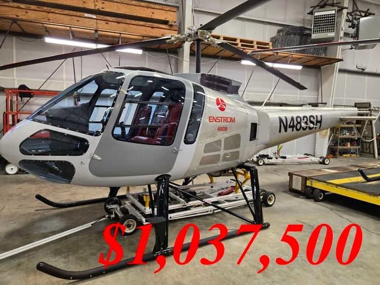 A light grey Enstrom 480B helicopter with red interior seats, parked inside a hangar.