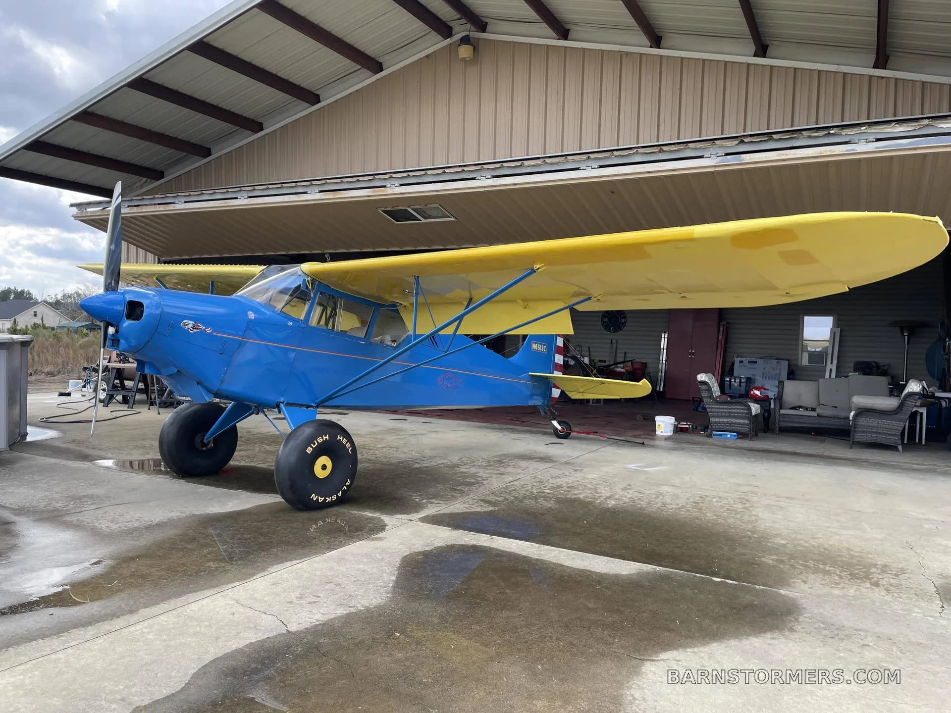 A blue and yellow high-wing aircraft with large tundra tires, registration N6513C, parked on a concrete pad outside an open hangar.