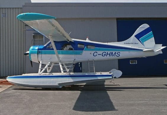 De Havilland DHC-2 Beaver floatplane in white and blue livery, parked near a hangar.
