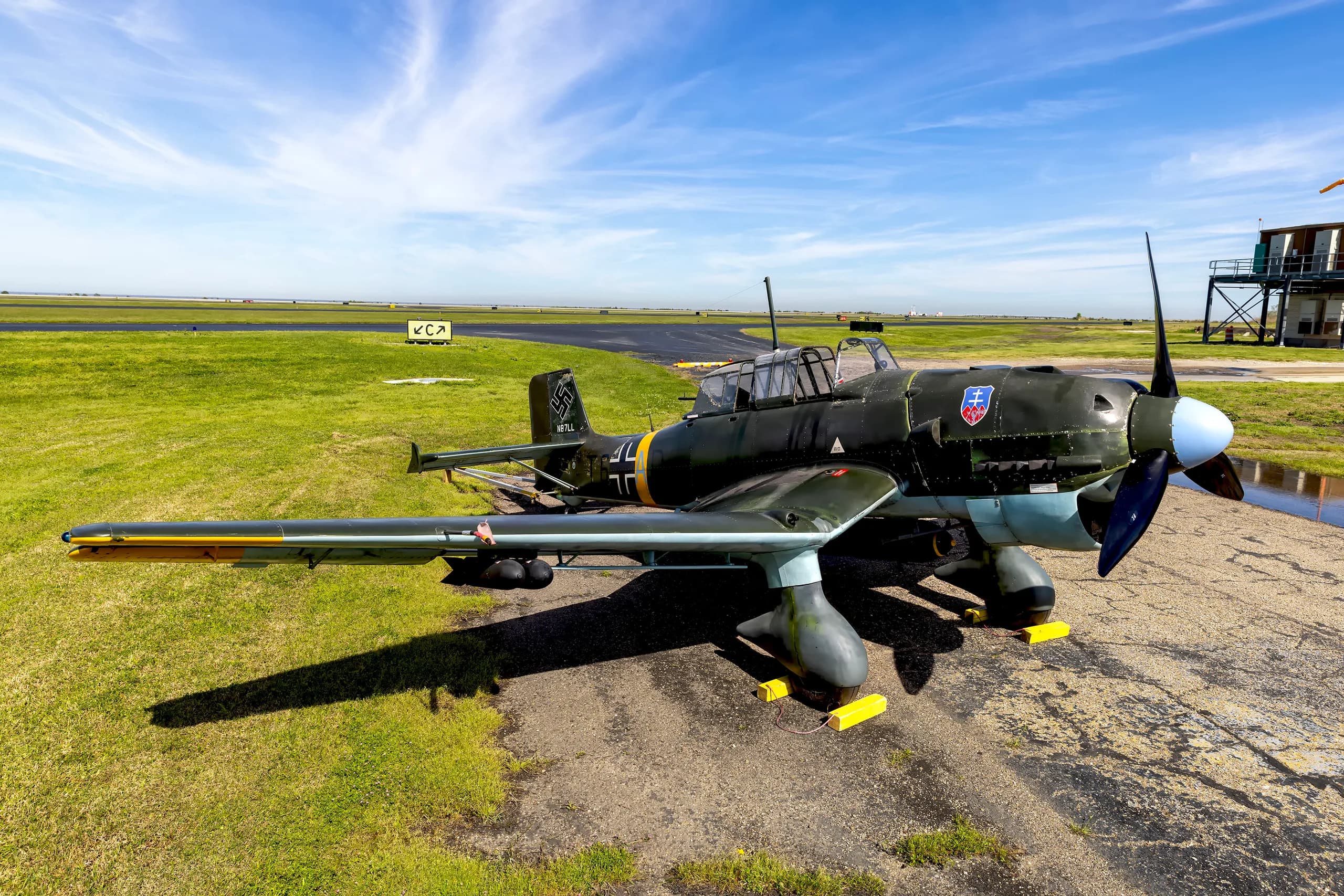 A three-quarter front view of a dark green Junkers Ju 87 Stuka on an airfield with a control tower in the background.