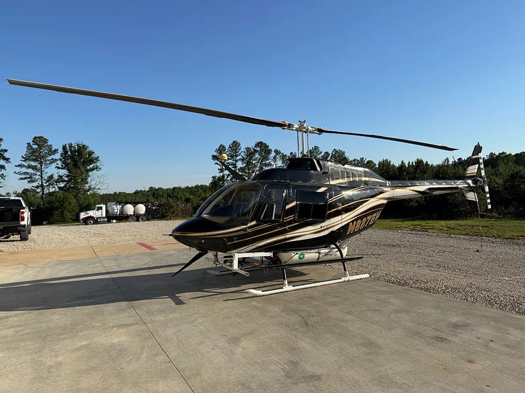 A black and gold helicopter, N887EH, sits on a concrete pad with trees and a truck in the background.