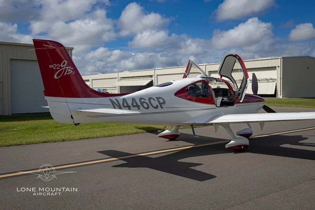 A red and white Cirrus SR22 aircraft (N446CP) with its doors open, viewed from the rear-left, parked on an asphalt ramp.