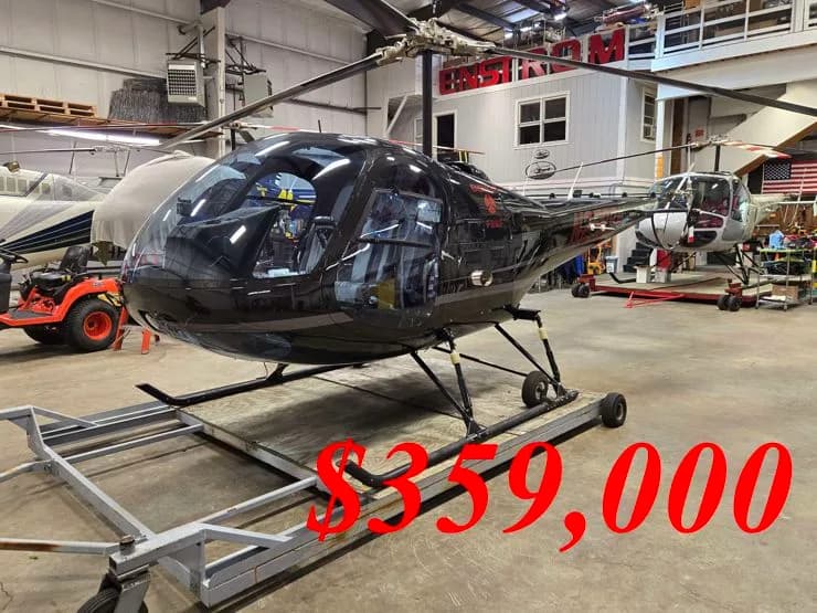 A black Robinson R44 helicopter on a dolly in a hangar, with other aircraft and equipment in the background.