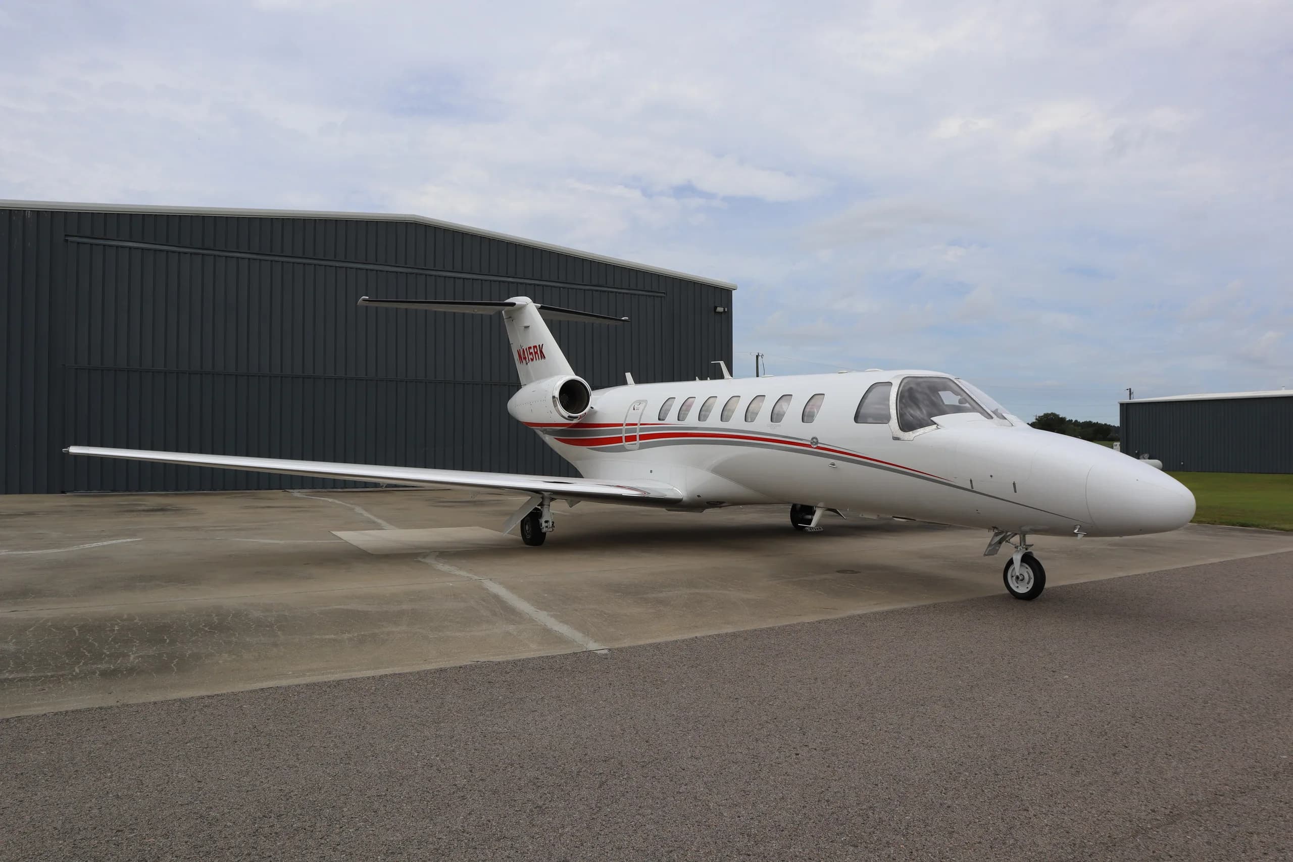 A white Cessna Citation business jet with red and grey stripes, registration N415RK, parked on an tarmac in front of a grey hangar.