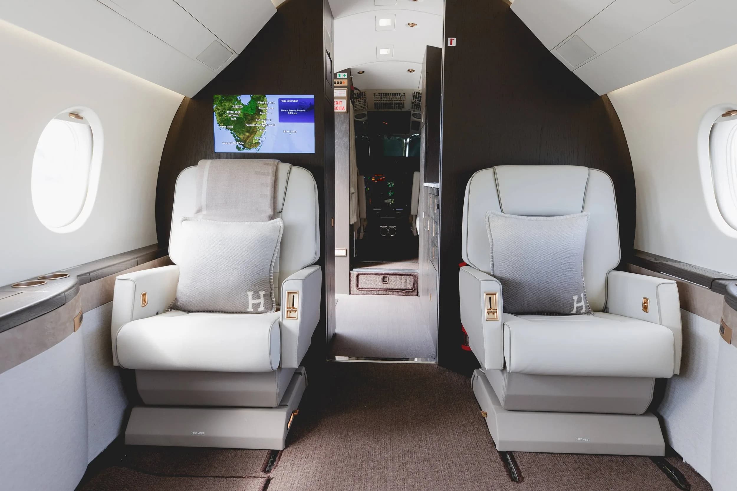 Two white leather seats with "H" monogrammed pillows facing a screen displaying a map, with the cockpit door visible beyond.