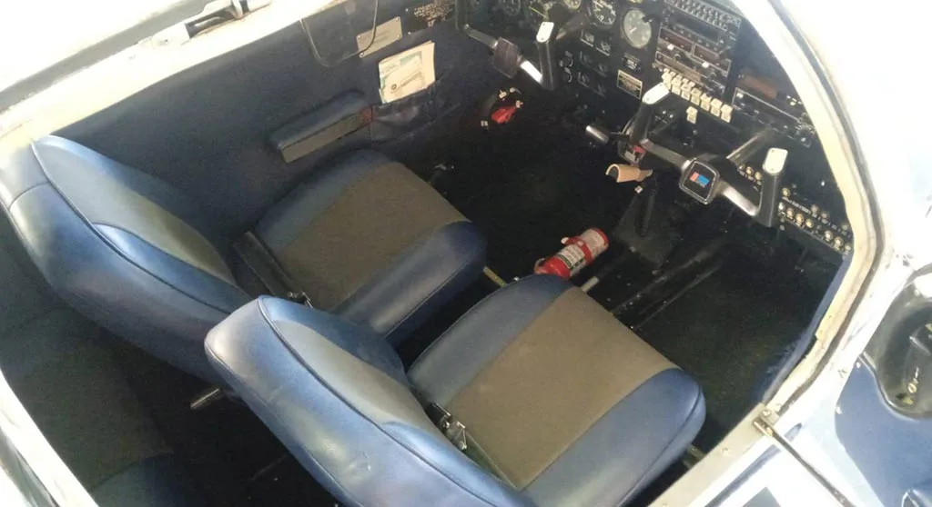 The front cabin seats of a small aircraft, upholstered in blue and grey, with a partial view of the cockpit.