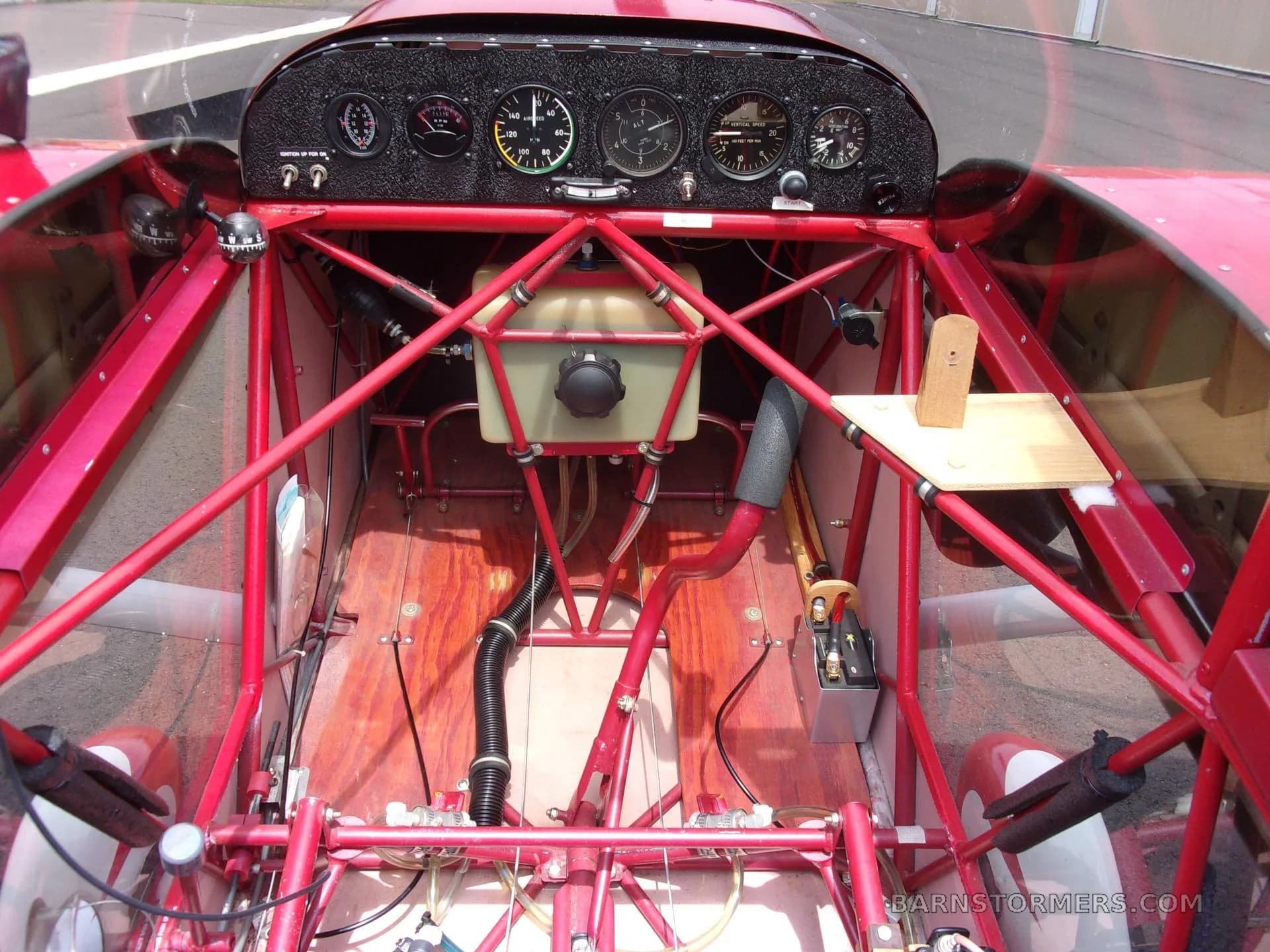 The open cockpit of a red aircraft, showing the instrument panel, control stick, and internal structural tubing.