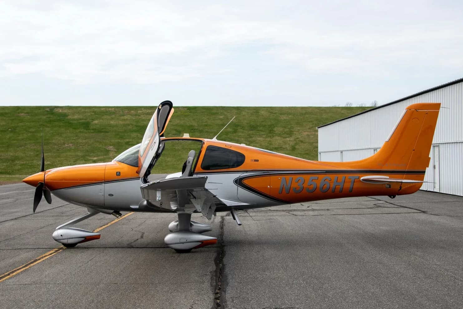 A side view of a metallic orange and silver Cirrus SR22 aircraft (N356HT) with its gull-wing doors open, parked on an asphalt ramp.