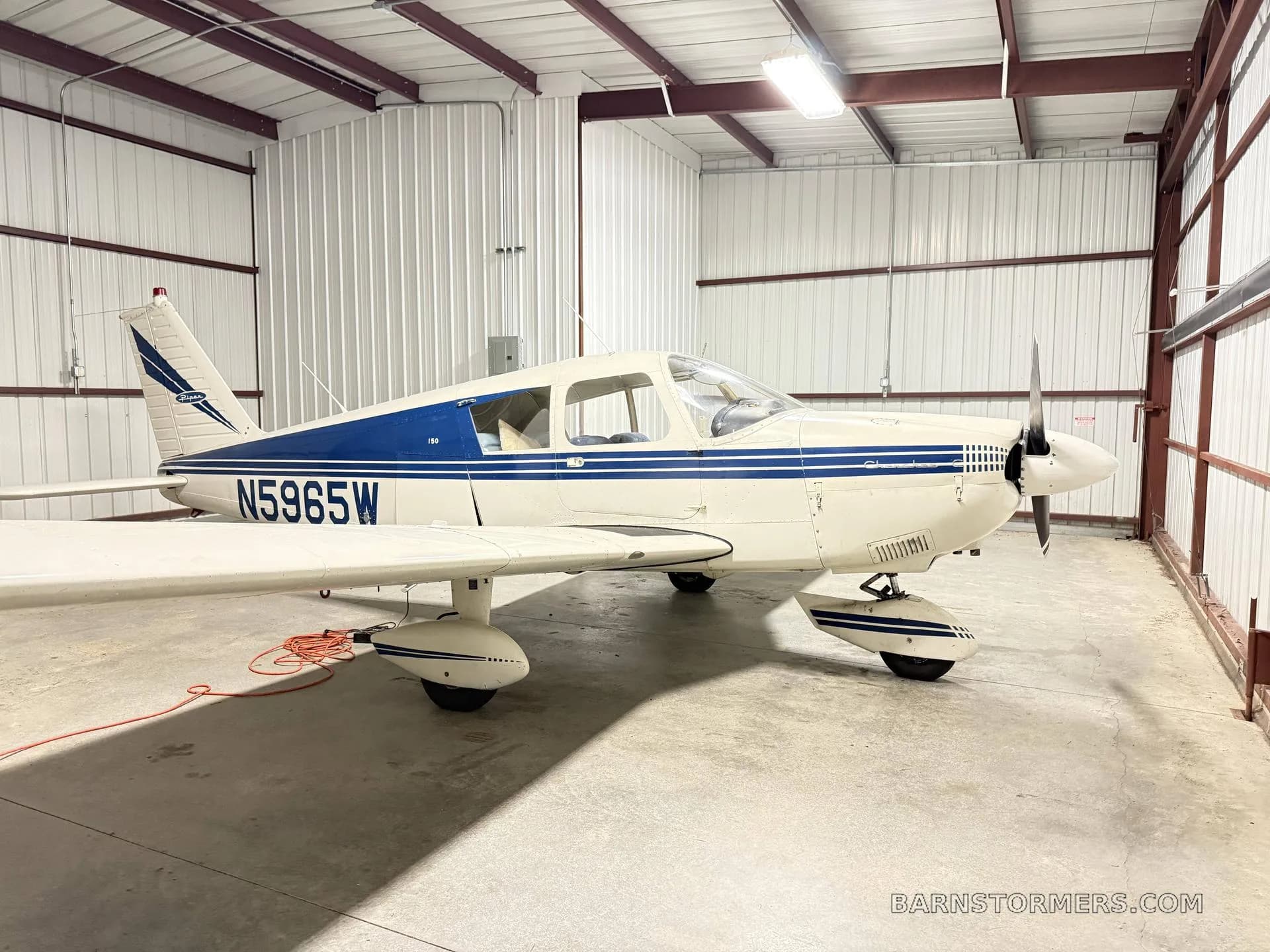 A white Piper Cherokee 150 aircraft with blue stripes and tail number N5965W parked inside a hangar.