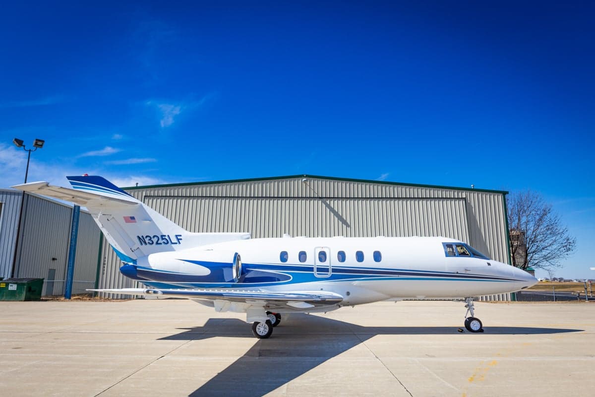 A white and blue jet sits on the tarmac in front of a hangar on a sunny day.