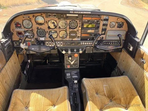 The cockpit of a small aircraft, featuring a brown instrument panel with various gauges, radios, and two control yokes, with tan upholstered seats.