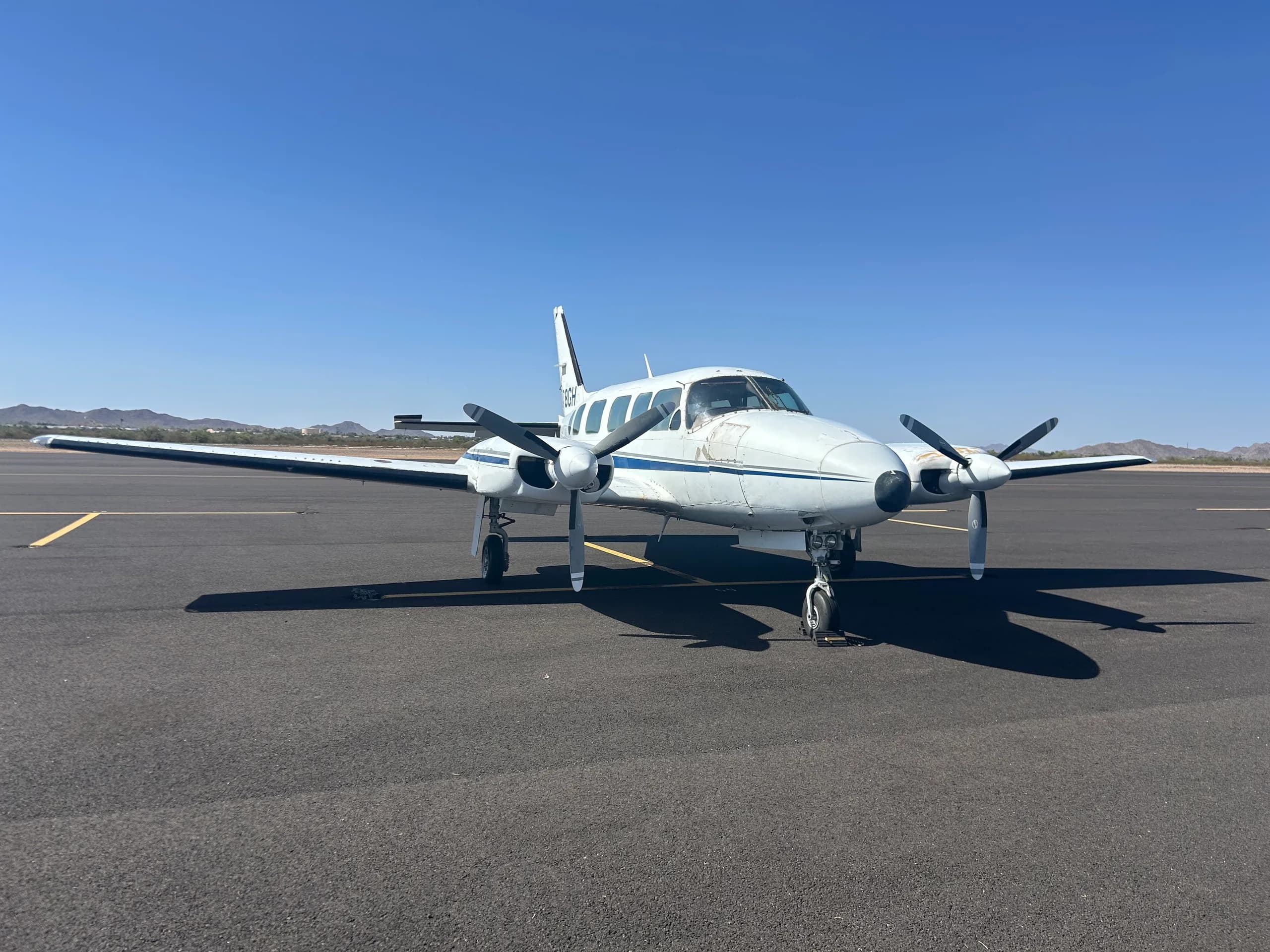 A white twin-engine aircraft with blue and grey stripes, registration N78GH, parked on an asphalt tarmac under a clear blue sky.