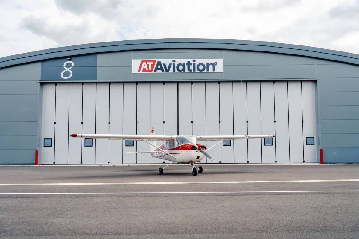 A white and red Cessna 150 G-ASMS parked directly in front of a grey AT Aviation hangar.