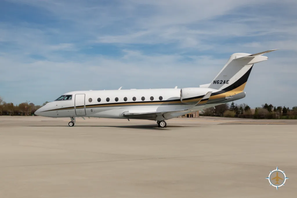 A white and gold business jet (N62AE) parked on a concrete tarmac under a blue sky.