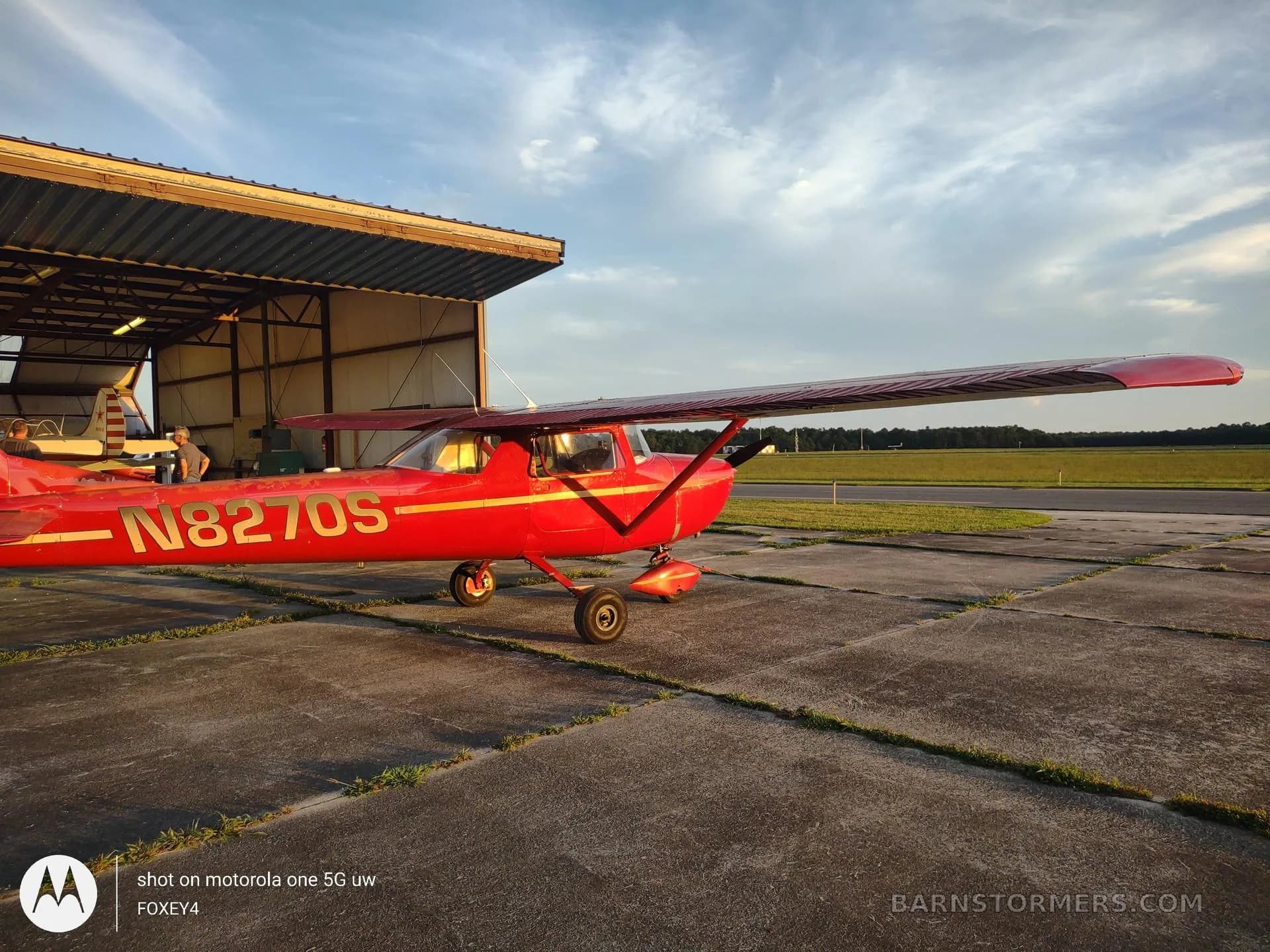 A red Cessna 150 or similar light aircraft with registration N8270S is parked on a concrete apron outside a hangar at sunset.