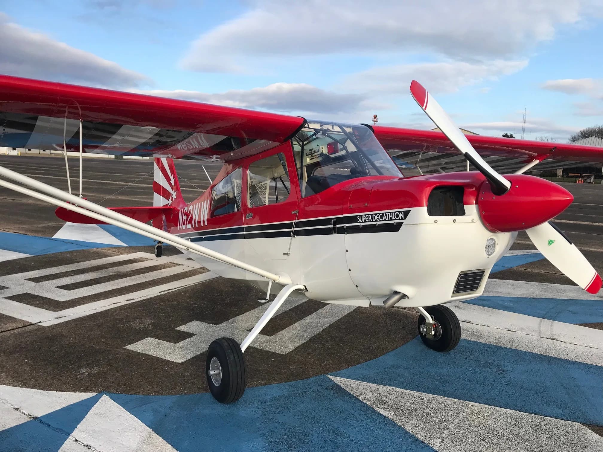 A red and white Super Decathlon aircraft, registration N62WW, parked on a blue and white marked tarmac.