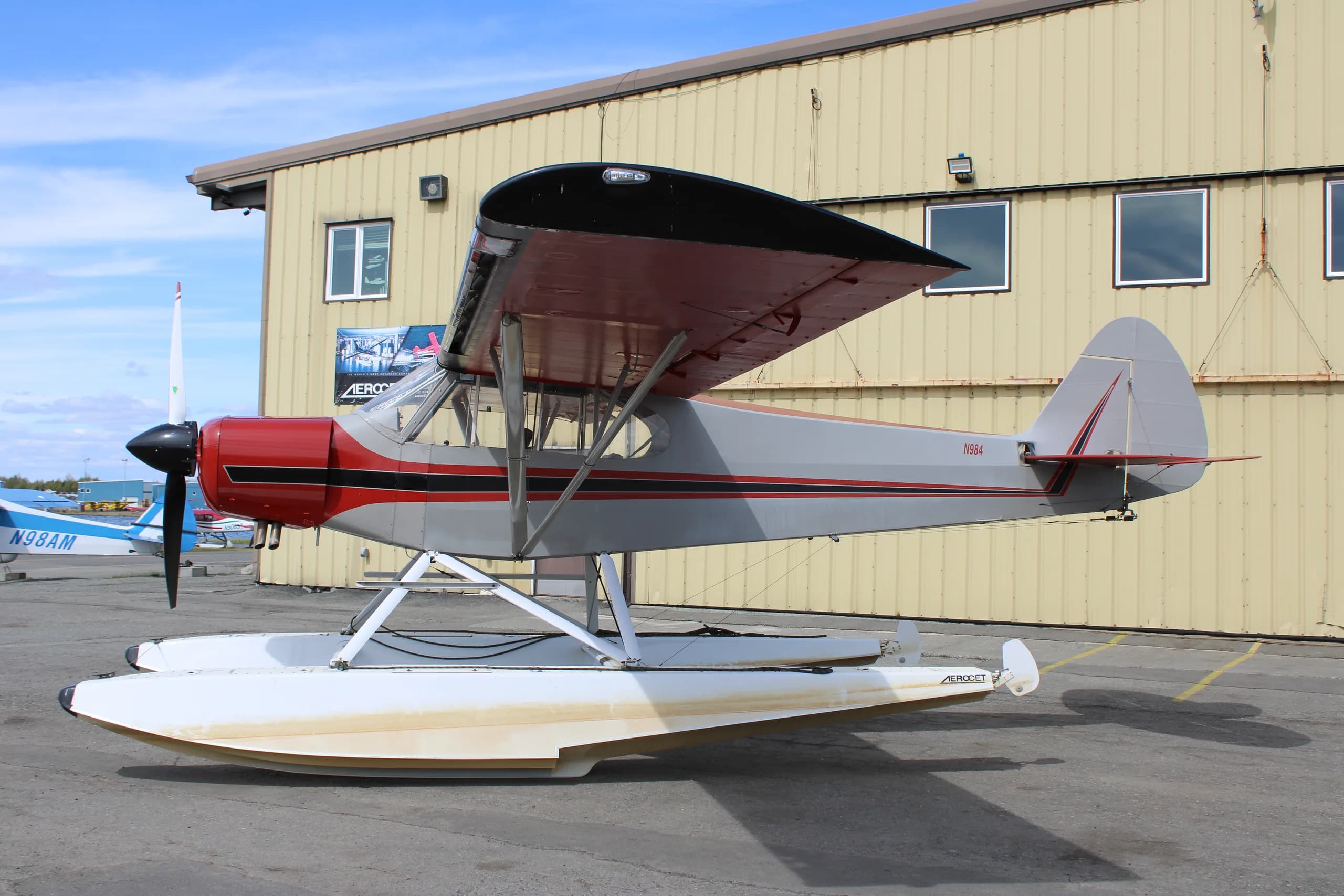 A red and silver float plane is parked in front of a hangar, side view.