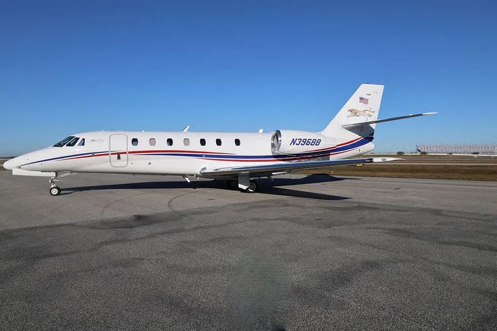 A three-quarter side view of the white Cessna Citation business jet N396BB with red and blue stripes, parked on an airfield under a clear blue sky.
