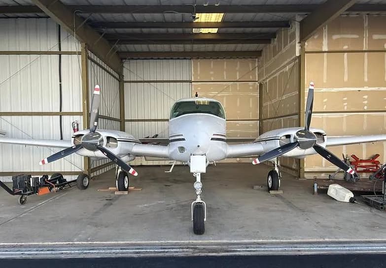 A white twin-engine propeller aircraft parked inside a hangar, viewed from the front.