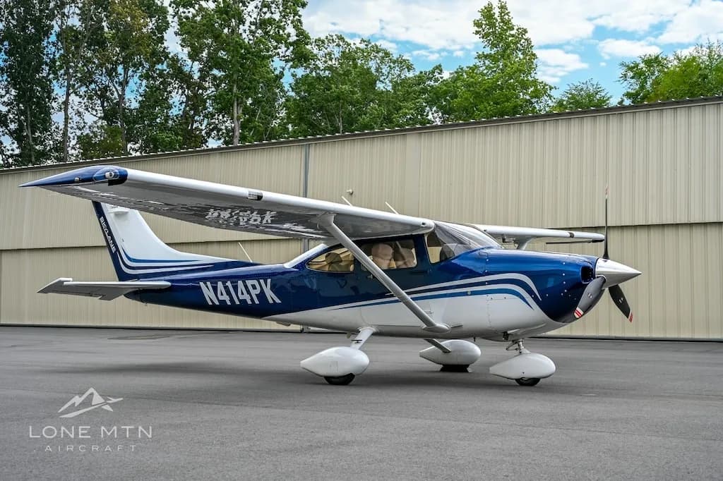 A blue and white Cessna Skylane N414PK parked on an asphalt ramp with hangars and trees in the background.