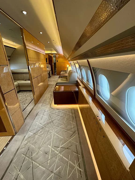 A luxurious aircraft interior features a patterned carpet, wood paneling, and individual seats along a brightly lit aisle.
