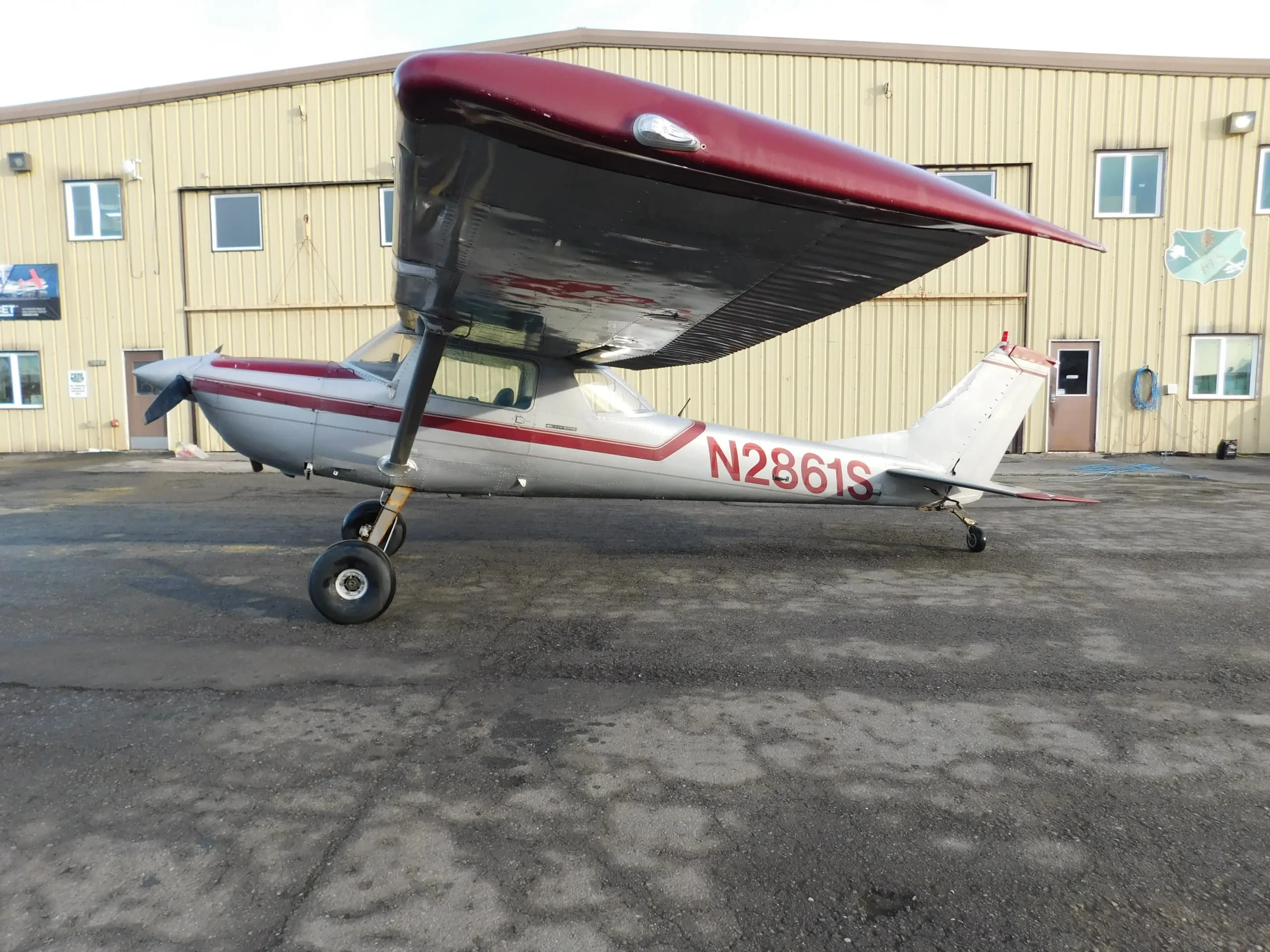 A silver and red high-wing single-engine aircraft with large tires is parked on an asphalt apron in front of a light yellow building.