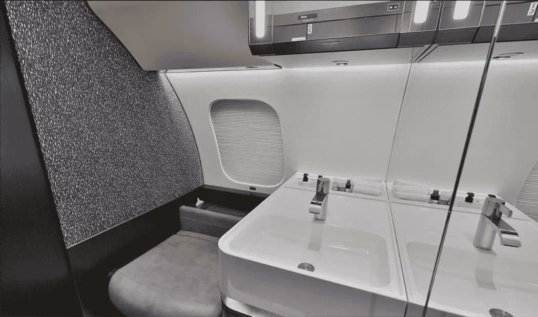 A modern aircraft lavatory with a white sink, mirror, and textured grey wall paneling.
