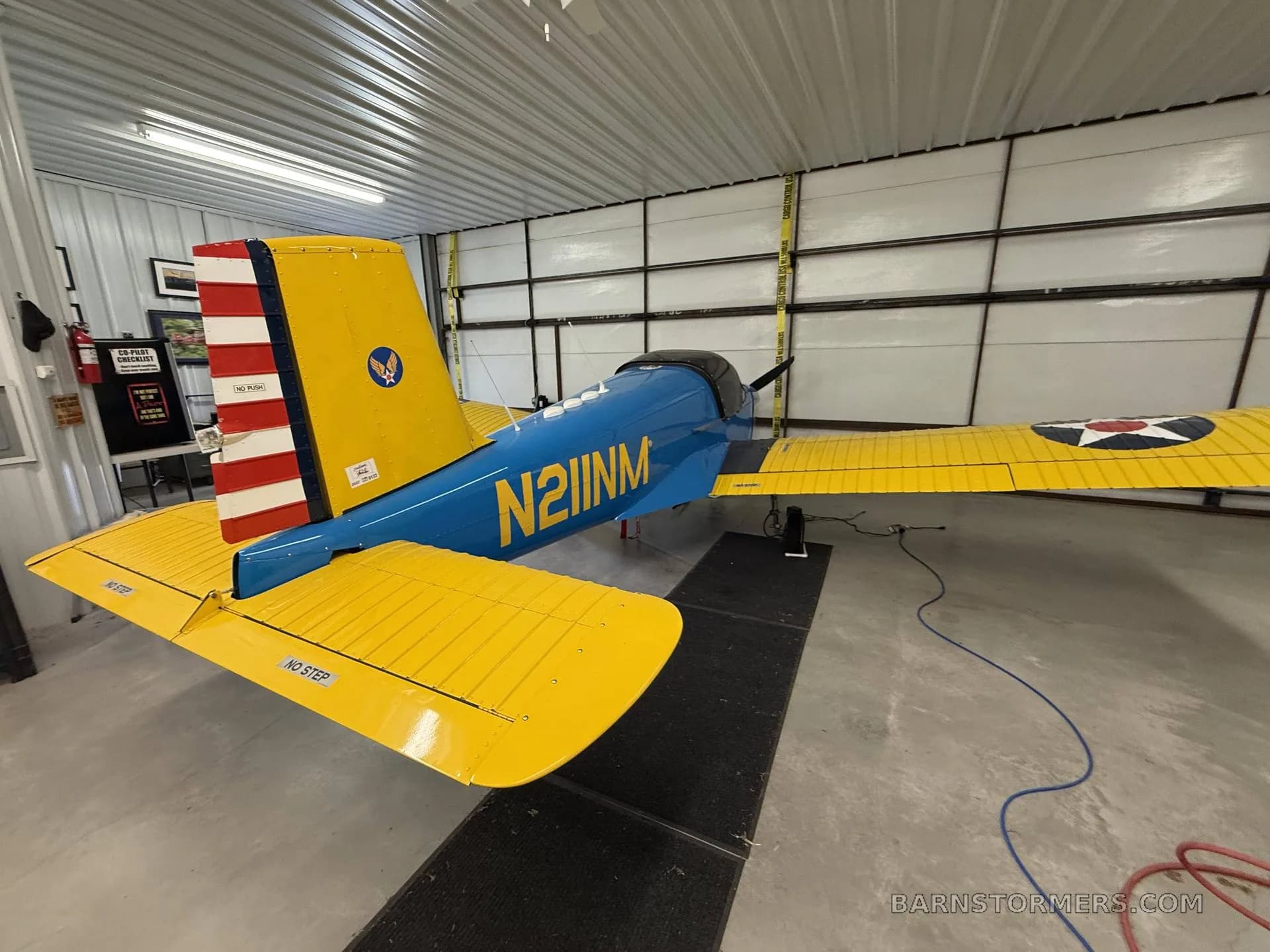 Rear view of a blue and yellow single-engine aircraft with 'N211NM' on the fuselage and a US star insignia on the wing, in a hangar.