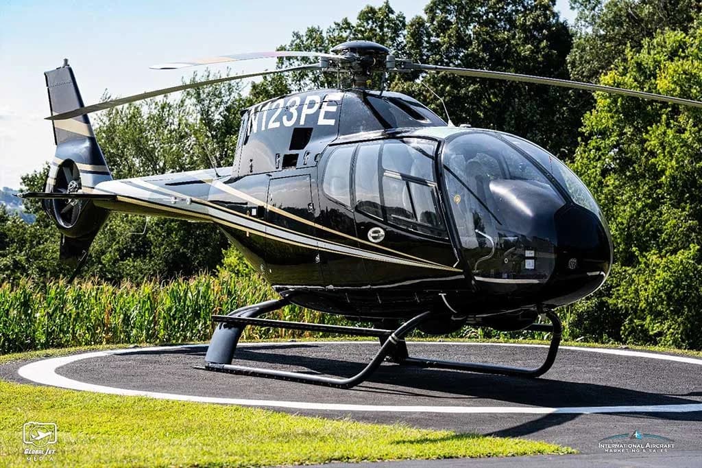 A black Eurocopter AS350 helicopter (N123PE) with silver stripes on a helipad surrounded by trees.