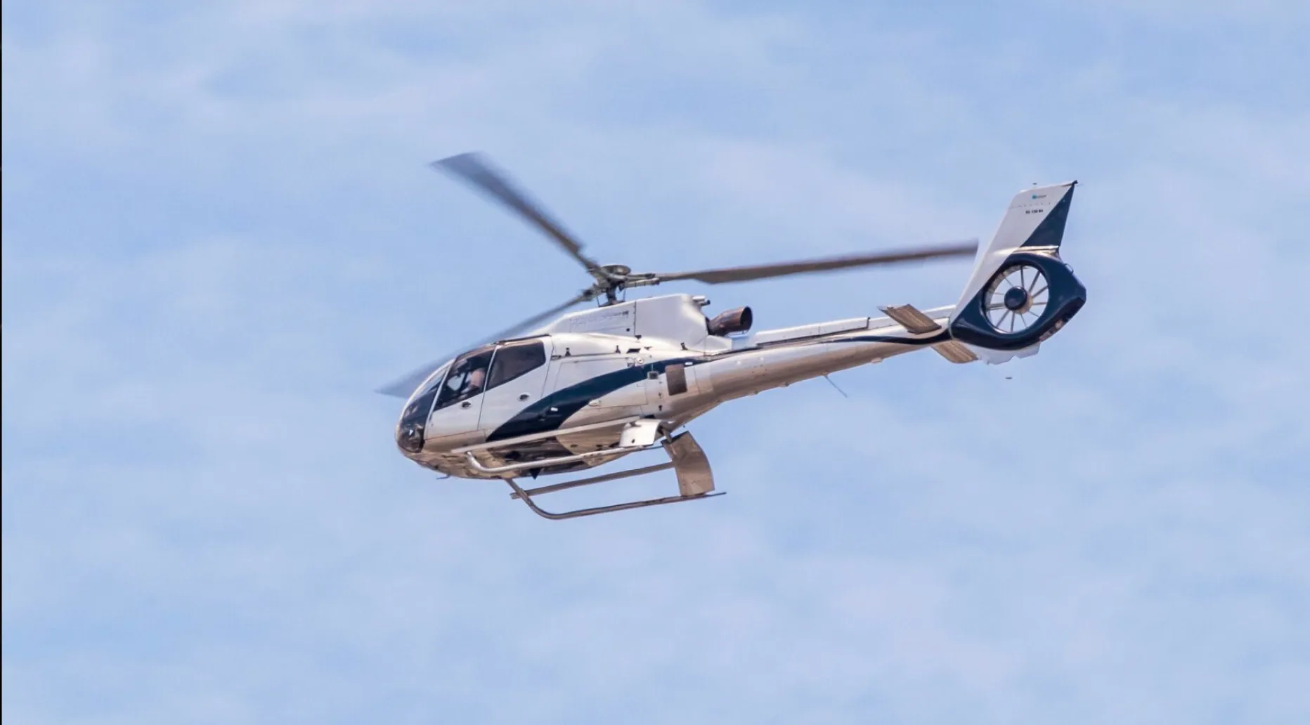 A silver and black Eurocopter EC130 B4 helicopter in flight against a blue sky.