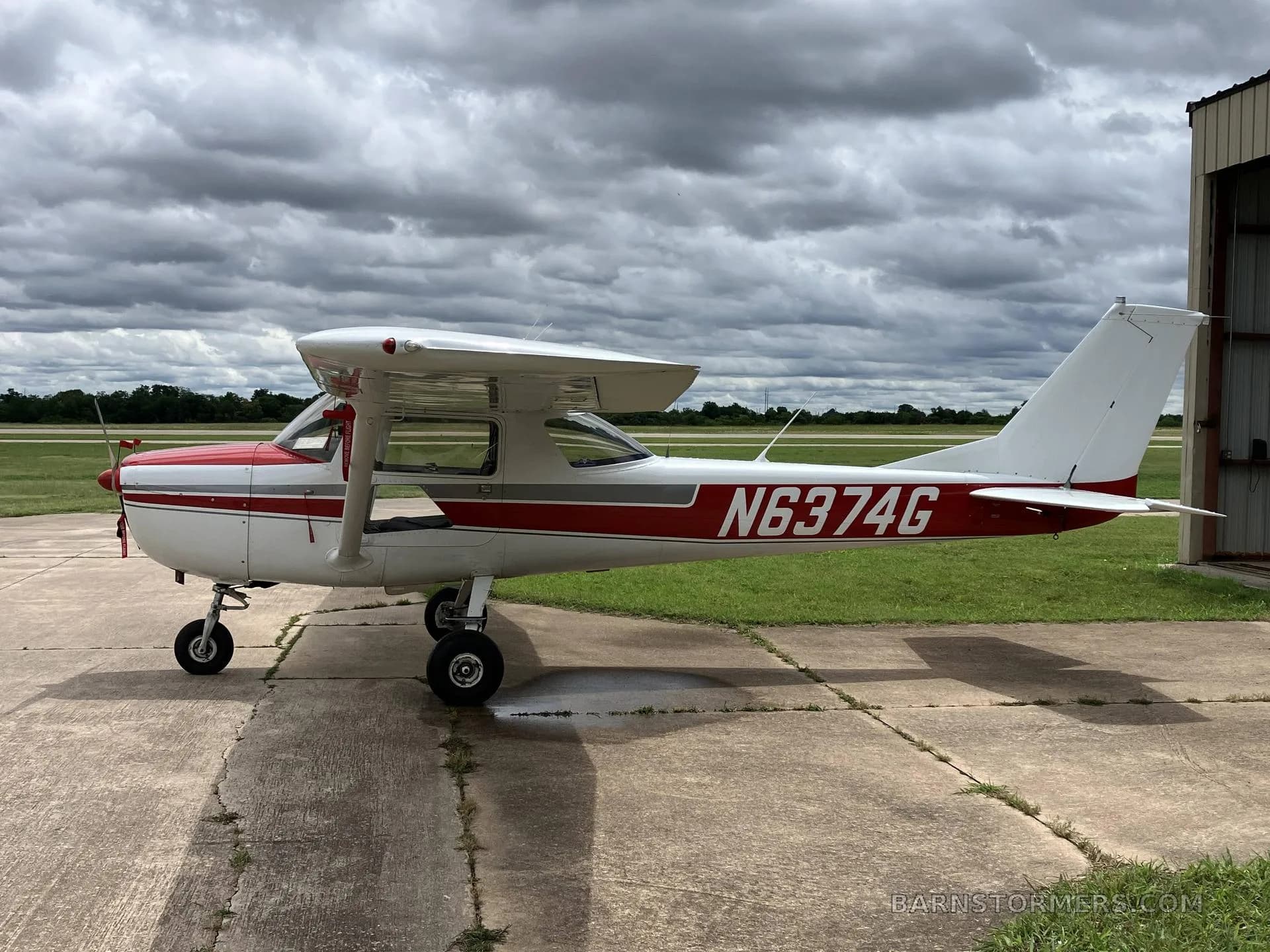 A white and red Cessna 172 aircraft with registration N6374G parked on a concrete pad next to a grassy field under a cloudy sky.
