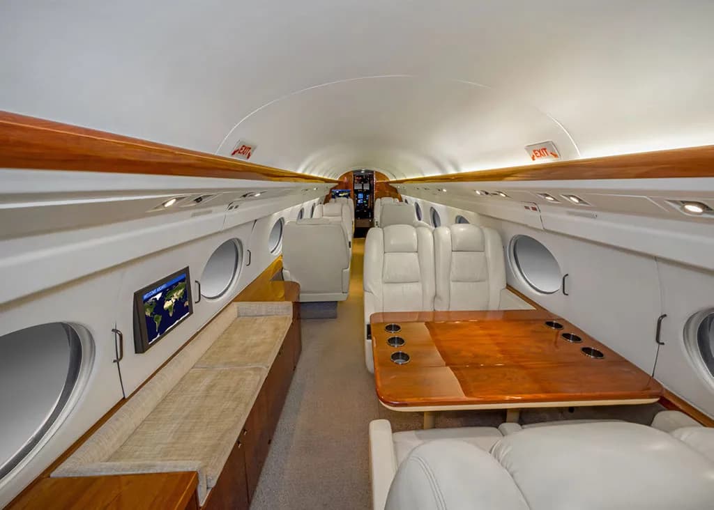 The interior cabin of a private jet features a large wooden table with seating, a long bench, and a monitor displaying a world map.