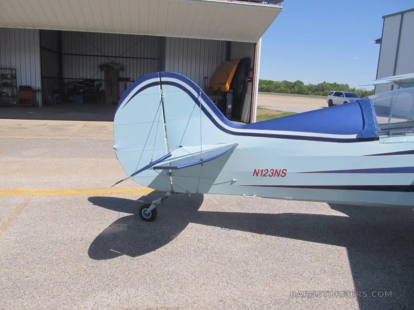 The tail section of a blue and light blue biplane, showing the N123NS registration, is parked near an open hangar.
