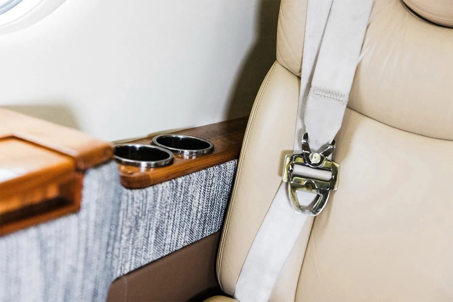 A close-up of a beige leather seat with a seatbelt and a wooden armrest with cup holders in the private jet's cabin.