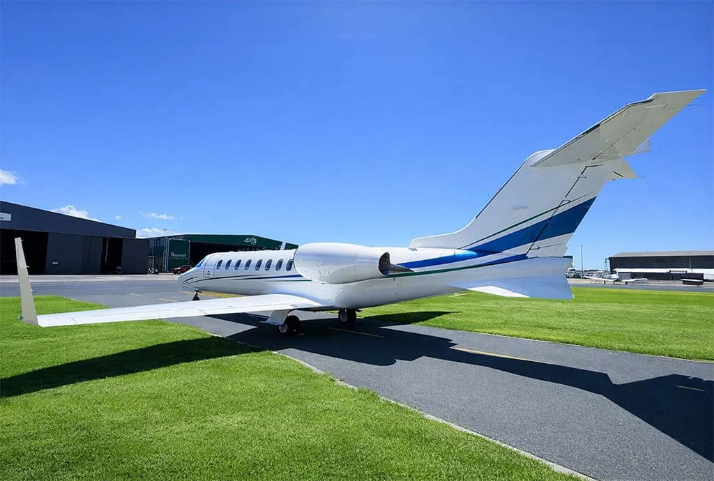 A white business jet with blue and green stripes is parked on an airport tarmac, viewed from the rear-side, with hangars under a clear blue sky.