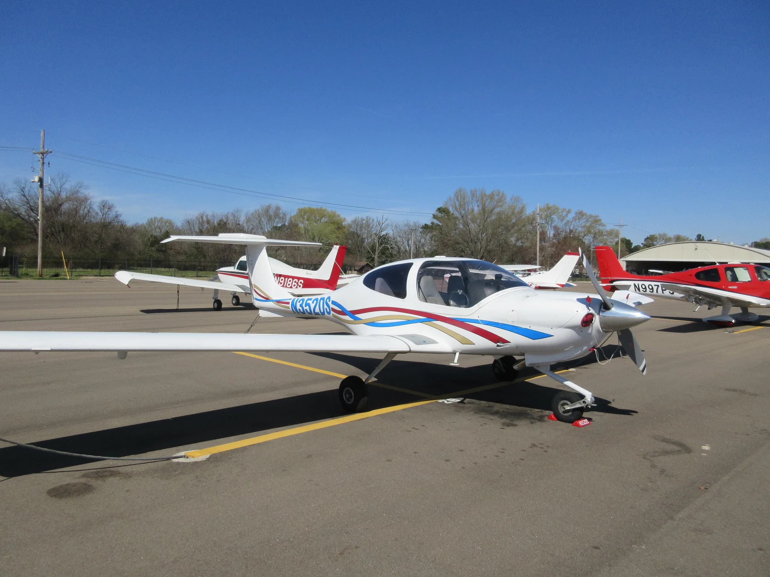 A white Diamond DA40 aircraft with blue, red, and gold stripes, registration N352DS, parked on an airport tarmac with other aircraft.