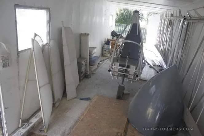 A workshop with a metal aircraft frame, a grey nose cone, and white panels leaning against a wall.