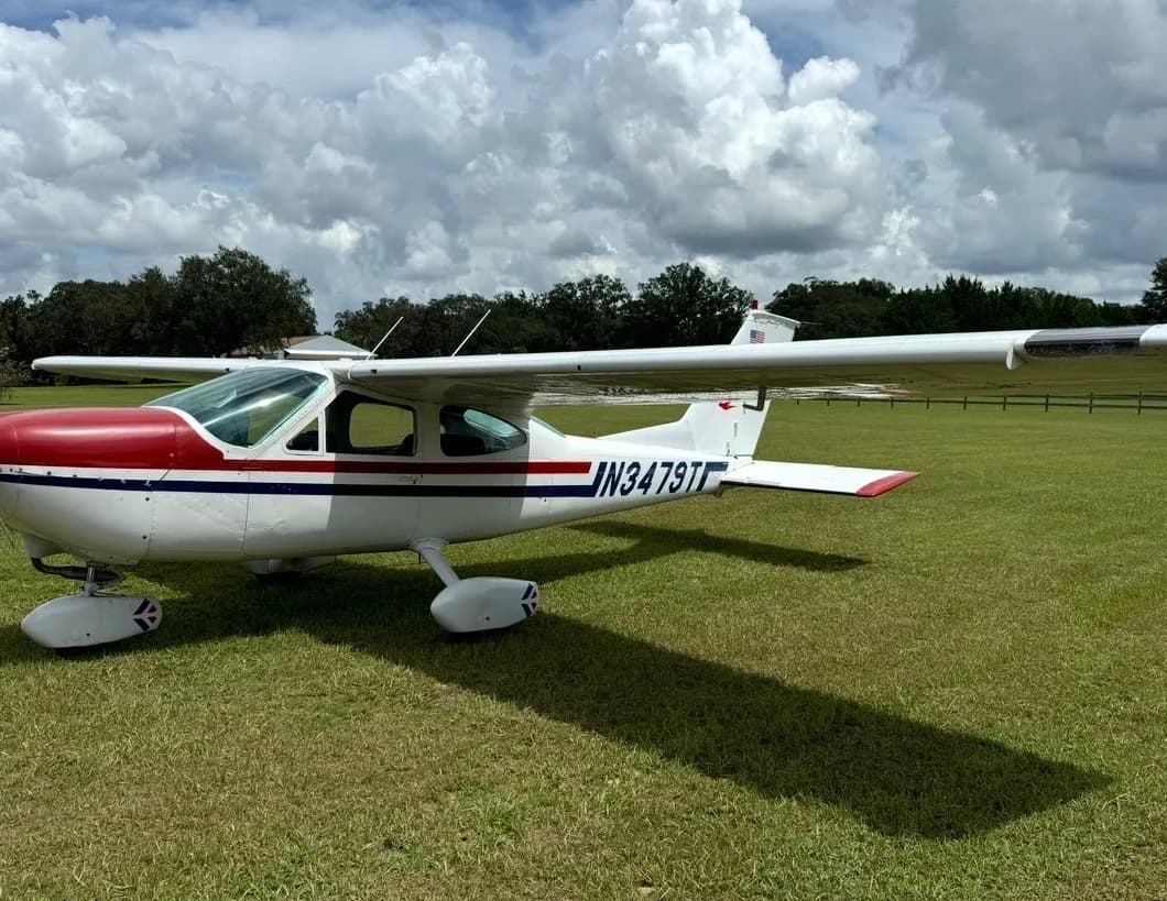 A white Cessna 182 with red and blue stripes, registration N3479T, parked on a green grassy field under a cloudy sky.