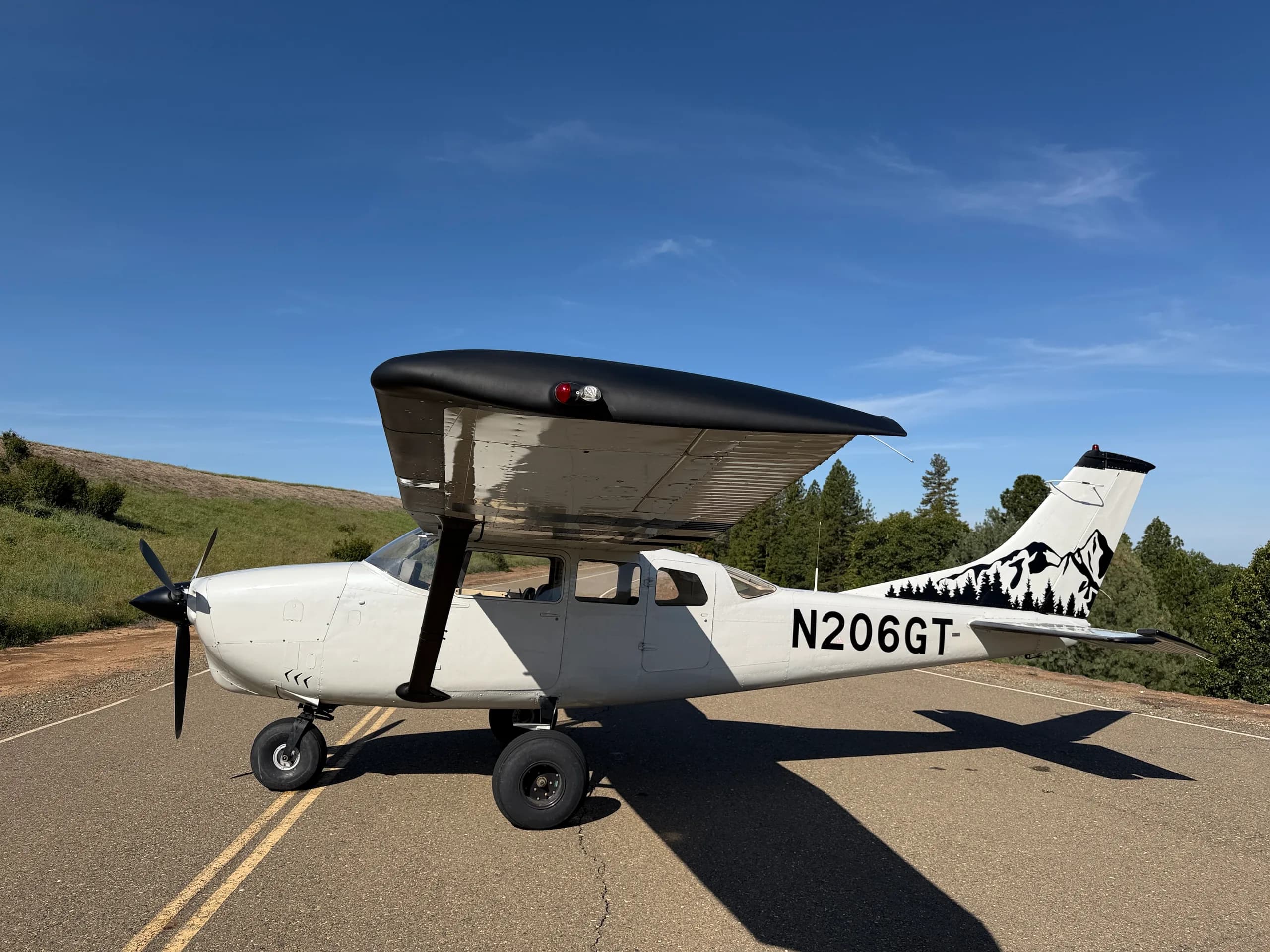 A white Cessna 206 with a black top wing and a mountain/forest decal on the tail is parked on an asphalt strip under a clear blue sky.