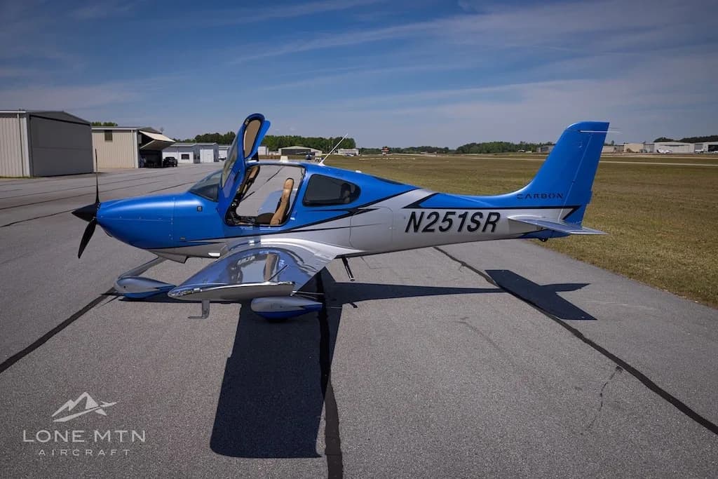A side view of the blue and silver Cirrus SR20 aircraft (N251SR) with open doors, parked on an airfield near hangars.