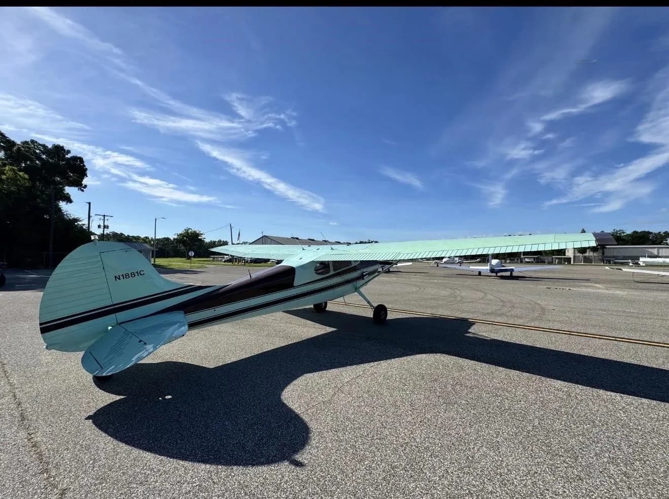 A rear-side view of a brown and light green Cessna 170B (N1881C) on an airport tarmac, casting a large shadow.