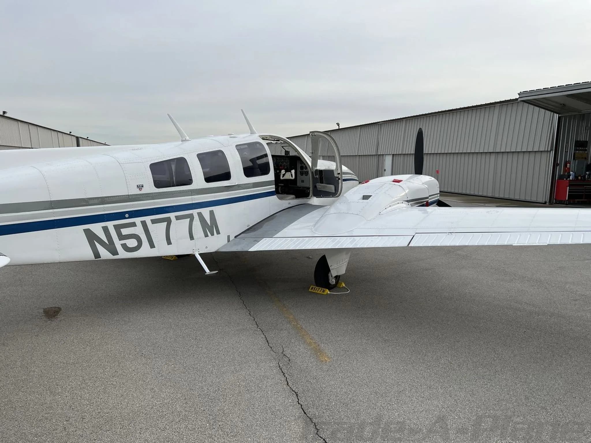 A side view of the white and blue Beechcraft Baron N5177M with its main cabin door open, parked on an airfield.