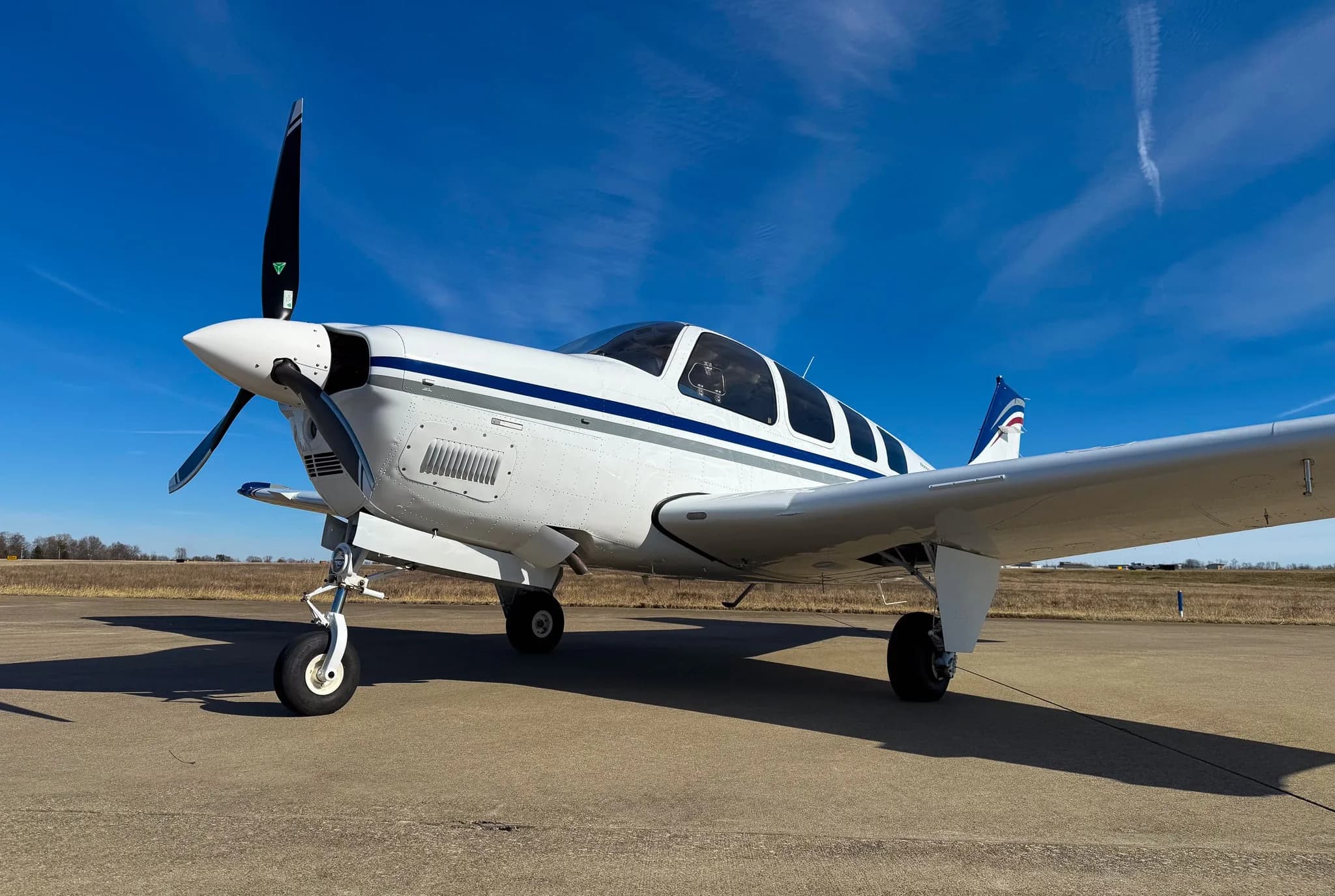 A low-angle front view of a white Beechcraft Bonanza G36 with blue and grey stripes, registration N7CM, parked on an airfield.