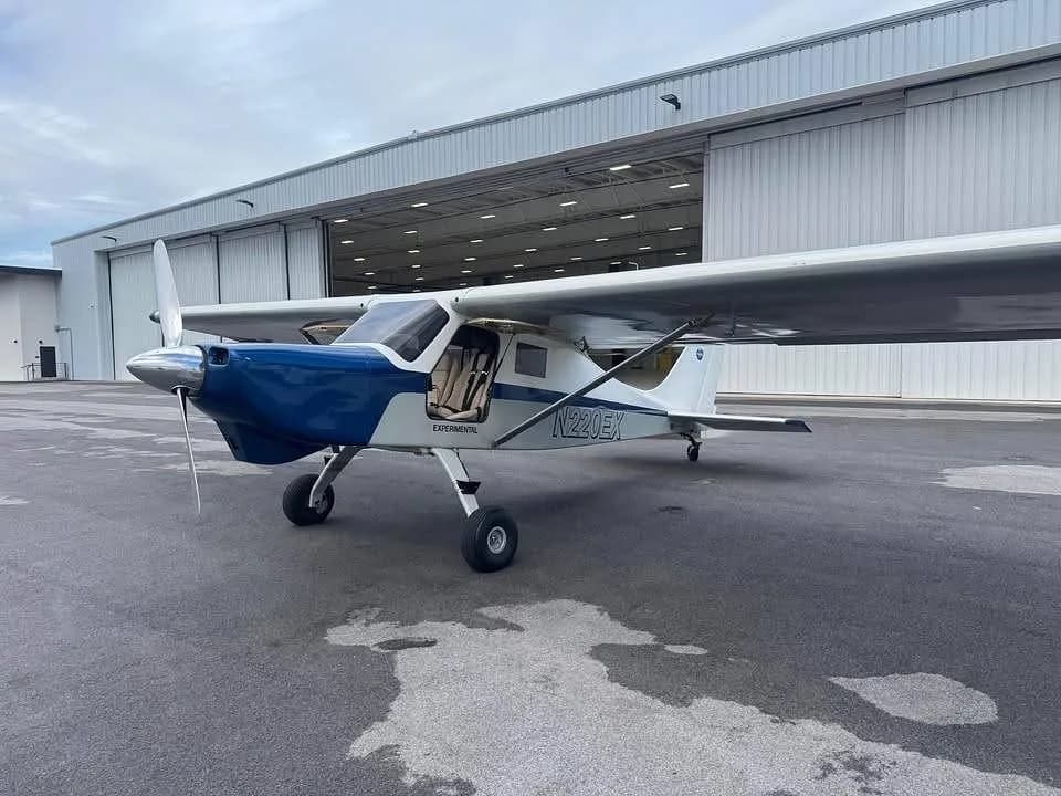 A white and blue experimental aircraft N220EX parked on a tarmac in front of an open hangar.