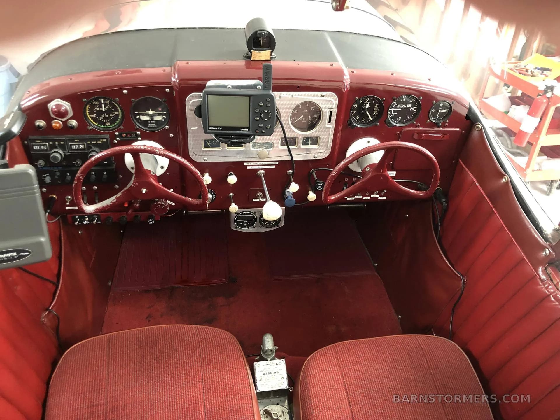 The cockpit of an aircraft features a red instrument panel, dual control yokes, and red upholstered seats.
