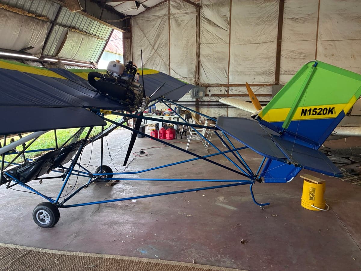 Rear-side view of a blue, green, and yellow ultralight aircraft with registration N1520K on the tail, parked in a hangar.