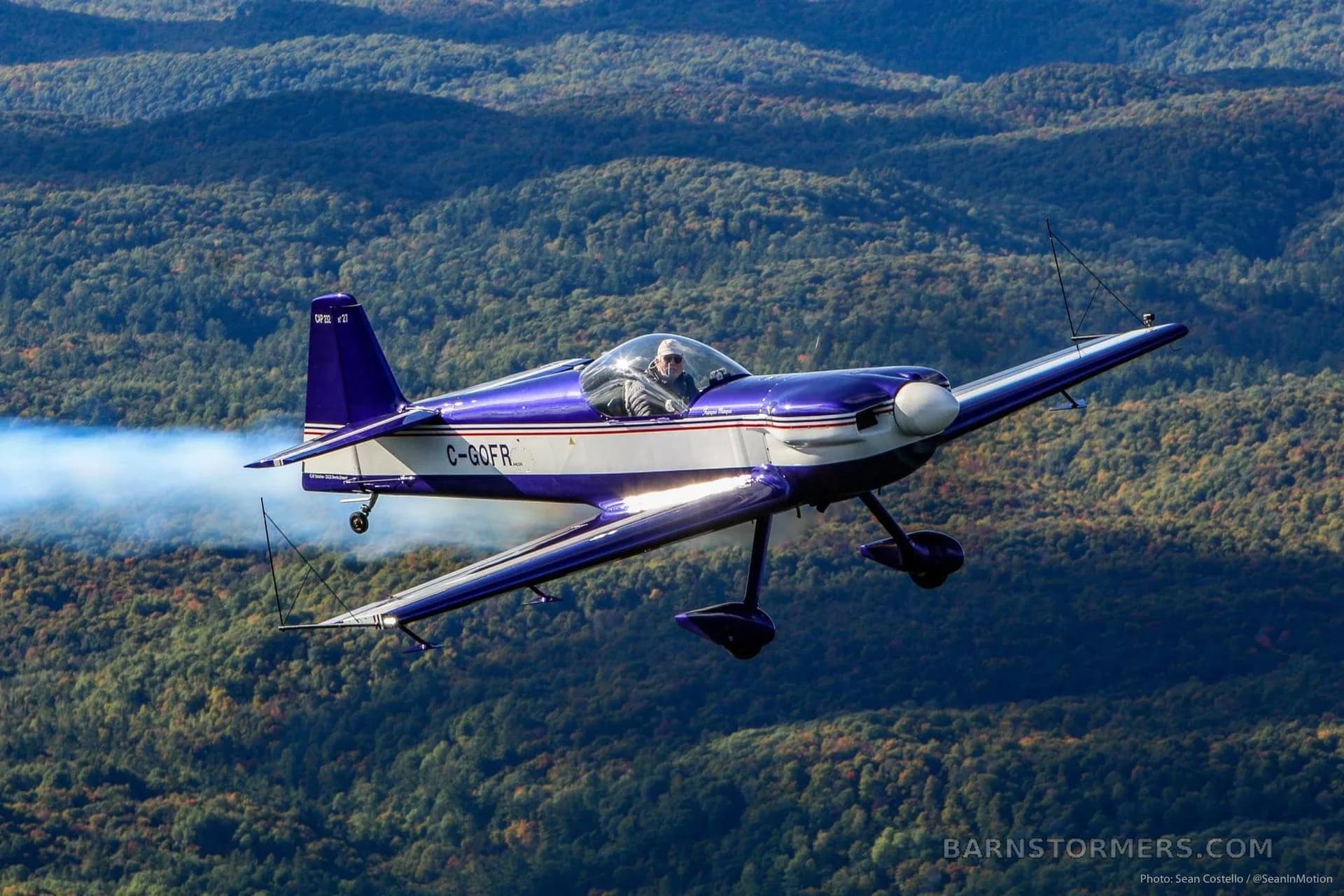 A blue and white CAP 10B aerobatic aircraft with registration C-GOFR flies over a forested landscape, trailing white smoke.